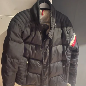 Svart pufferjacka från Moncler - Säljer en svart pufferjacka från Moncler med färgglada detaljer på ärmen. Jackan har en quiltad design och dragkedja framtill. Perfekt för kyligare dagar med sin stilrena look och varma material. Den är i begagnat skick, men fortfarande bra. 