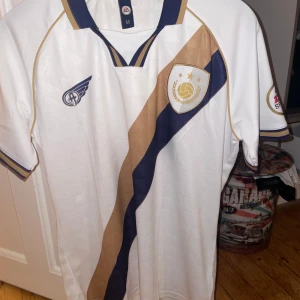 Vit fotbollströja från EA Sports - Säljer en vit fotbollströja från EA Sports med guld- och marinblå detaljer. Tröjan har korta ärmar och ett unikt mönster med ett emblem på bröstet. Perfekt för fotbollsfans som vill ha något speciellt i garderoben. Den är snålt använd och är i nyskick. Fläckfri