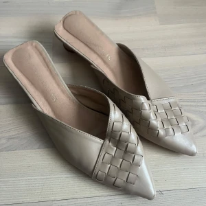 Beige klackskor med flätad detalj - Snygga beige klackskor med spetsig tå och flätad detalj framtill. Skorna har en låg klack och är perfekta för en stilren look. De är tillverkade i syntetmaterial och har en öppen häl för enkel påtagning.