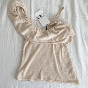 Beige topp från Zara - Säljer en söt beige topp från Zara med volangdetaljer och knytning i ryggen. Toppen har en offshoulder. Helt ny med lappar kvar, storlek S!
