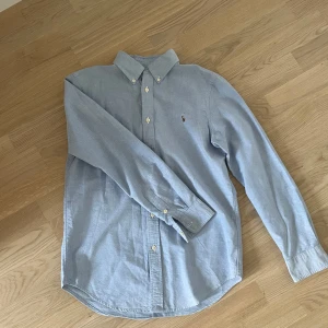 Ralph Lauren skjorta - Säljer en stilren blå skjorta från Ralph Lauren med button-down krage och broderad logga på bröstet. Skjortan har långa ärmar och knappar framtill. Perfekt för en klassisk look. Storleken är XS