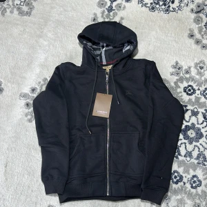 Svart hoodie från Burberry - Säljer en stilren svart hoodie från Burberry med dragkedja och klassiskt rutigt mönster i huvan. Perfekt för en avslappnad look. Den har långa ärmar och ribbade muddar vid ärmslut och nederkant.