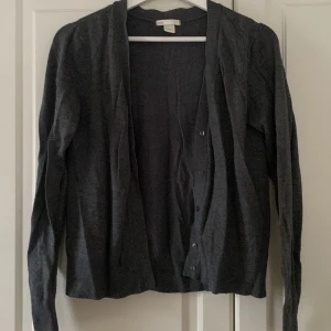Mörkgrå kofta från H&M Basic - Säljer en stilren mörkgrå kofta från H&M Basic. Den har långa ärmar och knappar framtill, perfekt för lager på lager-stil. Passar bra till både vardag och mer uppklädda tillfällen.