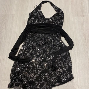 Svart glittrig klänning från Sisters Point - Snygg svart klänning från Sisters Point med glittrigt mönster och halterneck-design. Perfekt för en festlig kväll! Klänningen har en omlott-look och en v-ringad halslinje som ger en elegant touch.