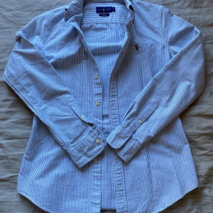 Blå och vit randig skjorta från Ralph Lauren - Säljer en stilren blå och vit randig skjorta från Ralph Lauren. Skjortan har långa ärmar och knappar framtill. Perfekt för en klassisk look. 🕶️