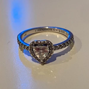 Silverring med hjärtformad sten från Pandora - Elegant silverring från Pandora med en gnistrande hjärtformad sten i centrum. Ringen är dekorerad med små stenar längs bandet som ger extra glans. Perfekt för att ge en romantisk touch till din stil.