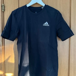 Svart t-shirt från Adidas - Snygg svart/mörkblå t-shirt från Adidas med deras klassiska logga på bröstet. Perfekt för en sportig look eller avslappnad stil. Kortärmad och tillverkad i mjukt material för extra komfort.