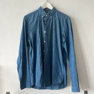 Blå jeansskjorta från Morris - Säljer en stilren blå jeansskjorta från Morris med klassisk krage och knappar framtill. Skjortan har långa ärmar och är tillverkad i ett autentiskt indigofärgat denimtyg. Perfekt för en avslappnad men ändå snygg look. Storlek M 39/40