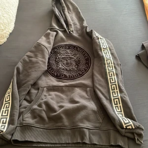 Grå hoodie från Glorious Gangsta - Säljer en grå hoodie från Glorious Gangsta med ett stort tryck på framsidan. Hoodien har en känguruficka och dekorativa band längs ärmarna. Perfekt för en avslappnad stil.