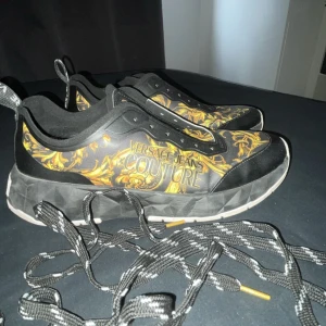 Svarta sneakers från Versace Jeans Couture - Snygga svarta sneakers med guldmönster från Versace Jeans Couture. Skorna har en unik design med ett iögonfallande mönster och logotyp på sidan. Perfekta för att ge din outfit en lyxig touch. Lite smutsiga bara.