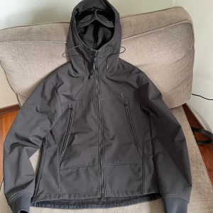 Svart Goggle Jacket från C.P. Company - Säljer en svart Goggle Jacket från C.P. Company. Jackan har en unik design med inbyggda glasögon i huvan och dragkedja framtill. Tillverkad i ett slitstarkt material med fleecefoder för extra värme. Perfekt för kyliga dagar.