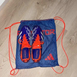 Adidas Predator fotbollsskor - Säljer ett par Adidas Predator elite fotbollsskor i en cool kombination av blått, vitt och orange. Skorna har snörning och är designade för att ge bra grepp på planen. Perfekta för fotbollsspelare som vill ha stil och funktionalitet.