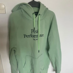Grön hoodie från Peak Performance - Säljer en snygg grön hoodie från Peak Performance med tryck framtill. Den har en klassisk känguruficka och justerbar huva med dragsko. Perfekt för en avslappnad stil.