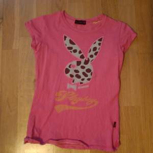 Säljer en rosa t-shirt från Playboy. T-shirten har ett  tryck av den ikoniska kaninen i ett mönster på framsidan och texten 'Playboy'på både fram och baksidan.