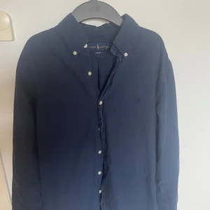 Mörkblå skjorta från Ralph Lauren - Snygg mörkblå skjorta från Ralph Lauren i slim fit. Skjortan har knappar framtill och en klassisk krage. Perfekt för en stilren look. Storleken är 175 men passar S/M 