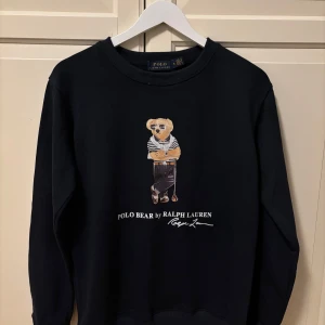 Svart tröja med Polo Bear från Ralph Lauren - Säljer en svart tröja från Ralph Lauren med det ikoniska Polo Bear-motivet på framsidan. Tröjan är tillverkad i 100% bomull och har en klassisk rund halsringning. Perfekt för dig som älskar stilrena och bekväma plagg!
