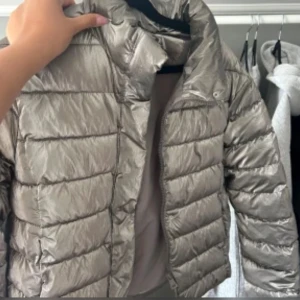 Silverfärgad dunjacka - Snygg och glansig silverfärgad dunjacka med dragkedja. Perfekt för kyliga dagar med sin quiltade design som håller värmen. Jackan har en hög krage och långa ärmar för extra skydd mot kylan.