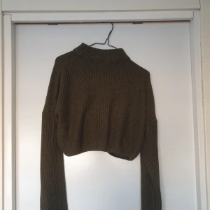 Olivgrön stickad croppad tröja - Säljer en olivgrön stickad tröja med croppad passform. Tröjan har långa ärmar och en ribbad krage som ger en stilren look. Perfekt för lager-på-lager under höst och vinter. Har en liten lös tråd, se bild