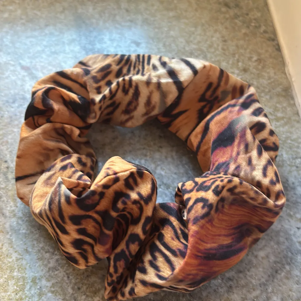 Snygg scrunchie med leopardmönster i bruna och svarta nyanser. Perfekt för att ge din frisyr en trendig touch. Tillverkad i ett mjukt och elastiskt material som sitter bekvämt i håret.. Asusteet.