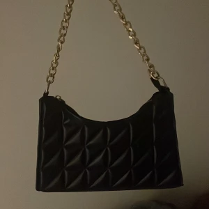 Svart quiltad axelväska med kedja - Snygg svart axelväska i quiltat mönster med en elegant guldfärgad kedja som axelrem. Väskan har en dragkedja upptill för säker förvaring. Perfekt för att ge en stilren touch till din outfit.
