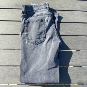 Grå jeans från SMOG - Säljer ett par grå jeans från SMOG i storlek W28 L30. De har en klassisk femficksdesign med knappgylf och bälteshällor. Perfekta för en stilren look.