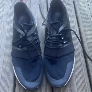 Svarta sneakers från Hugo Boss - Snygga svarta sneakers från Hugo Boss med stilren design. Skorna har snörning och en elastisk rem över foten för extra komfort. De har en vit sula och röda detaljer på hälen. Perfekta för en modern och avslappnad stil.
