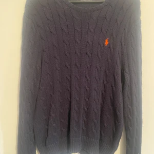 Mörkblå stickad tröja från Polo Ralph Lauren - Säljer en snygg mörkblå stickad tröja från Polo Ralph Lauren med kabelstickat mönster. Tröjan har en broderad logotyp i orange på bröstet och långa ärmar. Perfekt för kyliga dagar!