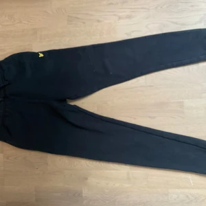 Svarta mjukisbyxor från Lyle & Scott Junior - Svarta mjukisbyxor från Lyle & Scott Junior med gul broderad logga framtill. Byxorna har resår i midjan och mudd vid bensluten. Perfekta för en avslappnad stil. Stl 12-13 år