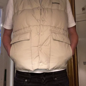 Beige dunväst från Marwin - Säljer en beige dunväst från Marwin med hög krage och knäppning framtill. Västen har två stora fickor med lock och tryckknappar. Perfekt för lager-på-lager och enkel att matcha med olika outfits.