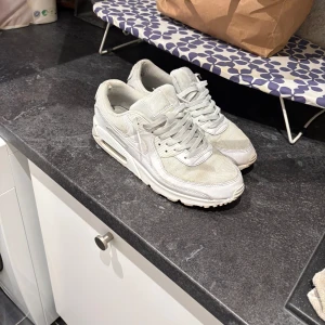 Vita Nike Air Max 90  - Säljer ett par klassiska vita Nike Air Max 90 sneakers med synlig luftbubbla i sulan och snörning. Skorna har en chunky siluett och är tillverkade i en mix av skinn och syntetmaterial. Perfekta för dig som gillar streetwear och sportig stil.