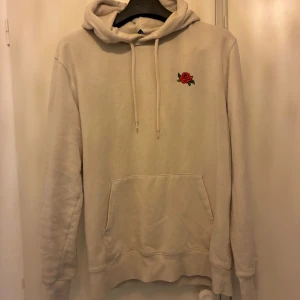 Beige hoodie med rosbrodyr - Säljer en beige hoodie med huva och dragsnören. På bröstet finns en liten broderad röd ros. Tröjan har en stor magficka och långa ärmar. Perfekt för en avslappnad stil.