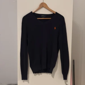 Mörkblå v-ringad tröja från Polo Ralph Lauren - Säljer en mörkblå v-ringad tröja från Polo Ralph Lauren i storlek S och är slim fit. Tröjan är i mjuk pima-bomull och har en orange broderad logga på bröstet. Perfekt för dig som gillar klassisk och stilren design.