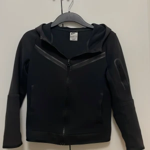 Nike tech fleece - Svart tech fleece från Nike med dragkedja framtill och huva. Tröjan har långa ärmar och diskreta detaljer på bröstet och ena ärmen. Perfekt för en sportig och avslappnad stil.