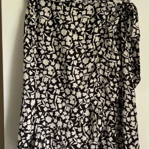 Svartvit blommig kjol från Amelie & Me - Säljer en blommig svartvit kjol från Amelie & Me med blixtlås och knytband i midjan. Perfekt i sommar. Mjukt skönt material. Aldrig använd 