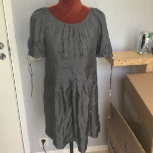 Grå klänning från Twist & Tango XS - Säljer en enkel och stilren grå klänning från Twist & Tango i storlek XS. Klänningen har korta puffärmar med knytband och veckad detalj framtill. Perfekt för dig som gillar minimalistisk stil. Grå klänning,bortklippta tvättråd  Gissningsvis polyester  Längd: 84 cm Armhåla till armhåla: 43 cm. Katter finns i hemmet!