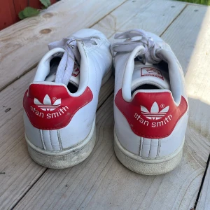 Adidas Stan Smith vita sneakers med röda detaljer - Klassiska vita Adidas Stan Smith sneakers med röda hälpartier. Skorna har en del repor, där av priset! Ingen sula då jag tagit ut och använt egna. Storlek 40 (jag har vanligtvis 39)