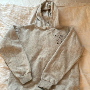 Ljusgrå hoodie från Gina Tricot - Mysig ljusgrå hoodie från Gina Tricot med huva och trycket 'Ma Chérie' samt en stjärna på bröstet. Perfekt för en avslappnad stil och enkel att matcha med det mesta. Mjuk bomullskänsla och ribbade muddar vid ärmslut och nederkant.