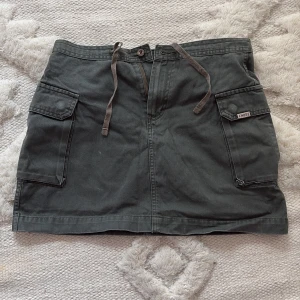 Grå minikjol med fickor från IKKS CORP - Säljer en grå / grön minikjol från IKKS CORP med stora fickor framtill och knytband i midjan. Kjolen har dragkedja och knappstängning samt detaljerade sömmar. Köpt i Köpenhamn. 