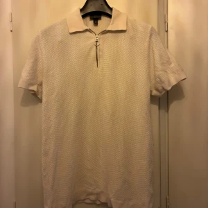 Vit pikétröja med dragkedja - Säljer en stilren vit pikétröja från H&M med korta ärmar och klassisk krage. Tröjan har en snygg dragkedja framtill istället för knappar och är tillverkad i ett strukturerat bomullsmaterial. Perfekt för en clean och fräsch look.
