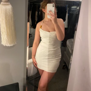 Vit halterneck klänning med tvinnade band - Säljer en snygg vit kortklänning med halterneck och tvinnade axelband med gulddetaljer. Klänningen har en figurnära passform och är perfekt för sommarens alla tillfällen. Enkel och stilren design som passar till mycket.