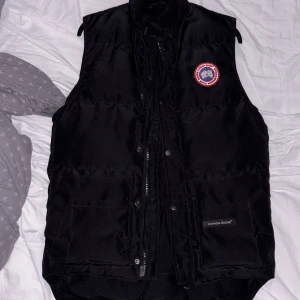 Svart dunväst från Canada Goose - Säljer en svart dunväst från Canada Goose med klassisk logotyp på bröstet och diskret varumärkeslapp vid fickan. Västen har dragkedja och knappar framtill och är perfekt för lager-på-lager-stil.