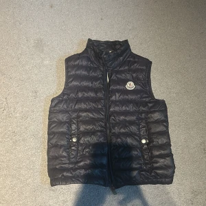Svart dunväst från Moncler - Snygg svart dunväst från Moncler med quiltad design och dragkedja framtill. Västen har två fickor med knappstängning och klassisk Moncler-logga på bröstet. Perfekt för lager-på-lager och stilren look.