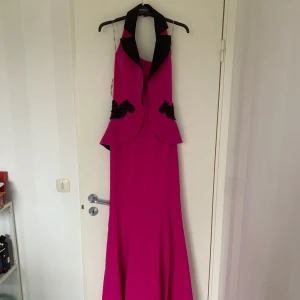 Cerise långklänning med svarta detaljer - Säljer en unik cerise långklänning med halterneck och svarta kontrastdetaljer. Klänningen har dekorativa svarta spetsapplikationer vid midjan och en elegant öppen rygg. Perfekt för dig som vill sticka ut på festen! Prislapp är på!!!