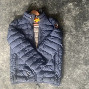 Blå dunjacka från Parajumpers - Säljer en blå dunjacka från Parajumpers i modellen Super Lightweight. Jackan har hög krage, dragkedja framtill och två sidofickor med dragkedja. Klassisk PJS-logga på ärmen och snygg quiltad design.