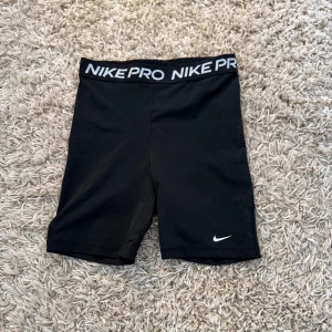 Svarta Träningsshorts från Nike Pro - Svarta tighta träningstights shorts från Nike Pro med vit logga på benet och bred resår i midjan med Nike Pro-tryck. Nick märket är lite slitet och shortsen är i storlek M