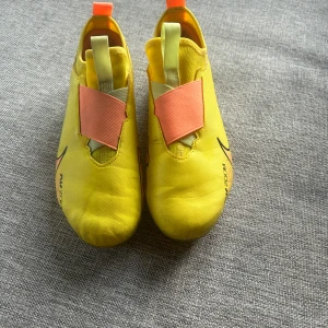 Nike Air Zoom fotbollsskor i gult - Säljer ett par gula Nike Air Zoom fotbollsskor med rosa och svarta detaljer. Skorna har en slimmad design, låg profil och elastiskt band över vristen. Perfekta för dig som vill sticka ut på planen! Från säljaren dom har tydlig användning på bilderna men ändå riktigt fräsch går däremot att negotiera sätter 500kr som pris men kom jätte gärna med pris förslag!🙌