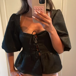 Svart glittrig blus med puffärmar och knytning - Säljer en svart glittrig blus med puffiga kortärmar och snygg knytning framtill. Blusen har en markerad midja och lätt volang nedtill. Perfekt för dig som vill sticka ut lite extra!