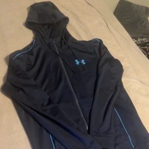 Under armour tracksuit  - Snygg svart träningsjacka med blå detaljer och dragkedja från Under Armour. Jackan har huva och lång ärm, perfekt för träning eller sportiga dagar. Under Armour-logga på bröstet och smidigt material. Och ett par matchande byxor från under armour med snygga detaljer. Koftan är storlek S byxorna är storlek S.