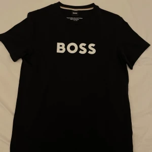 Svart t-shirt från BOSS med vit logga - Snygg svart t-shirt från BOSS med stor vit logga på bröstet. Tillverkad i bomull med regular fit och rund halsringning.Såklart äkta!✅Aldrig använd! Storlek XS! Skriv om ni har några funderingar!!