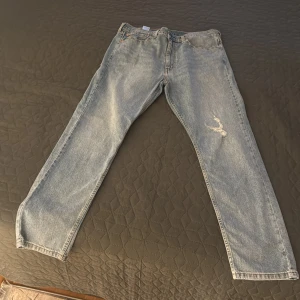 Levis 501 ljusblå jeans - Klassiska Levis 501 jeans i ljusblå denim med rak passform. Byxorna har en liten sliten detalj på höger lår och femficksdesign. Perfekta för en avslappnad stil och ikonisk look.